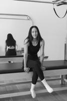 Black-and-white portrait of a woman in workout clothes sitting cross-legged on a Pilates reformer, smiling at the camera in a bright studio with mirrors and exercise equipment.