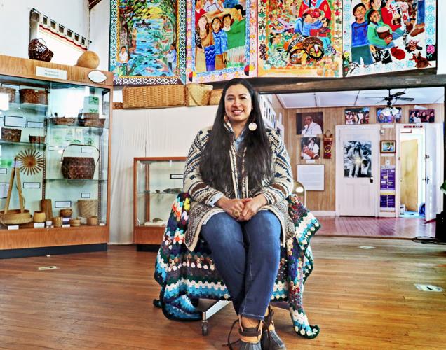 Speaker Silvermoon Mars LaRose with long, dark hair, wears a sweater and blue jeans. She sits in the center of the main display room of the Tomaquag Museum, surrounded by artifacts. There is a display case of woven baskets to her left; four colorful tapestries above her; and a smaller room with photographs on the wall to her right.