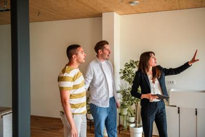 A Hispanic female real estate agent gestures informatively to men evaluating the spacious interior of a modern house.