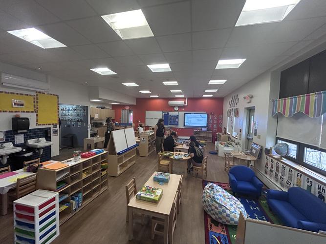 A bright classroom with children's tables, shelves of books, and a digital display screen.