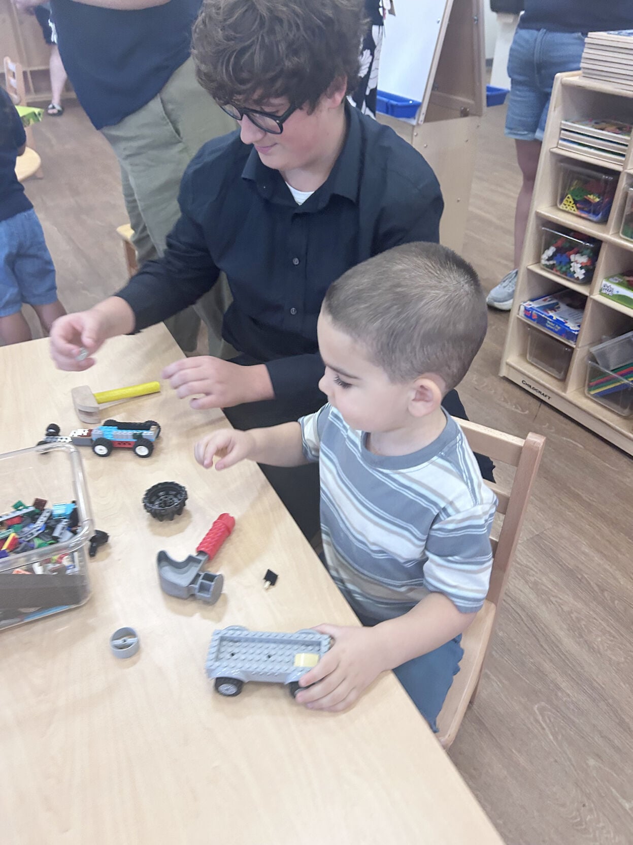 A young boy and an older teen build with toy bricks at a wooden table.
