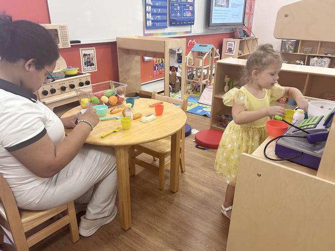 A child plays with a toy cash register while an adult sits at a round table with play food.