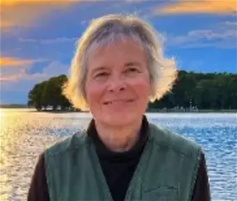 Woman with blond hair wearing a black mock shirt and green vest, standing in front of a lake