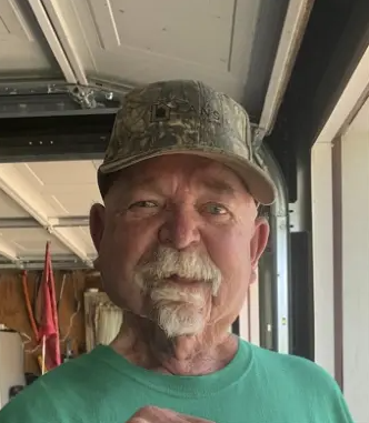 Man with mustache and goatee, wearing a green t-shirt & hunting cap standing in a garage with garage door open overhead.