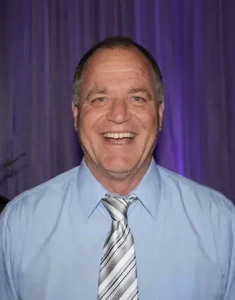 Man with short salt & pepper hair wearing a blue dress shirt, gray and black striped tie. Background purple curtain.