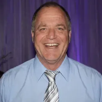 Man with short salt & pepper hair wearing a blue dress shirt, gray and black striped tie. Background purple curtain.