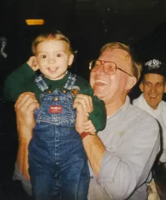Man wearing glasses & gray shirt, holding a toddler wearing blue coveralls