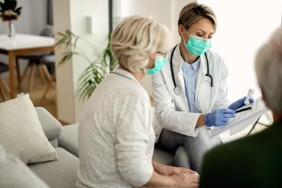 Female doctor and senior woman wearing protective face mask during home visit.