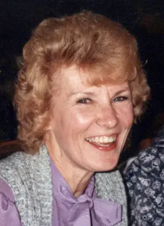 Woman with Red hair, wearing a purple top and gray vest, dark background