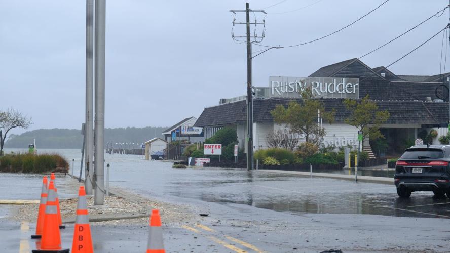 Orange traffic cones block a road covered with water. A building in the background has a sign that says Rusty Rudder.
