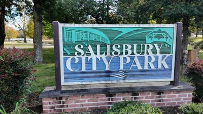 A sign reading Salisbury City Park with one of the park's walking trails, trees and a pedestrian bridge over water behind. 
