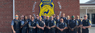Officers with the Laurel Police Department stand together in front of their logo on the side of a brick building.