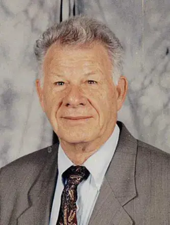 Man with white hair, wearing gray suit with a tie. Background is white with gray marbling wall. 