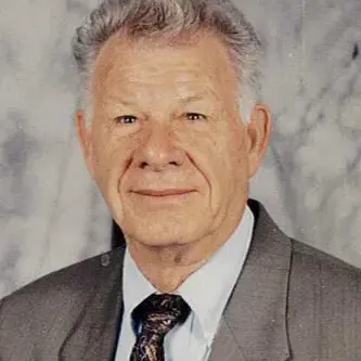 Man with white hair, wearing gray suit with a tie. Background is white with gray marbling wall. 