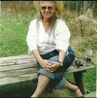 Woman with salt & pepper hair, wearing glasses, white 3/4 sleeve top, green caprice, and a black belt, sitting on a wooden bench outdoors.