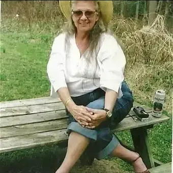 Woman with salt & pepper hair, wearing glasses, white 3/4 sleeve top, green caprice, and a black belt, sitting on a wooden bench outdoors.