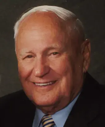 Man with white hair receding hair, wearing a black suit jacket, blue dress shirt. stripe tie, dark background.