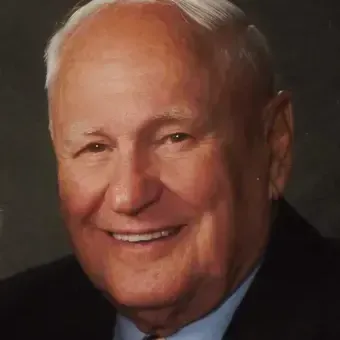 Man with white hair receding hair, wearing a black suit jacket, blue dress shirt. stripe tie, dark background.