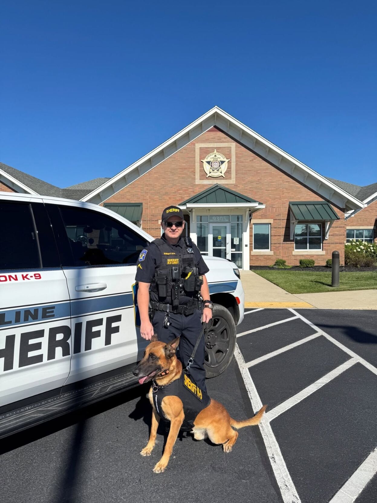 A sheriff's deputy stands next to a k9 wearing a vest.