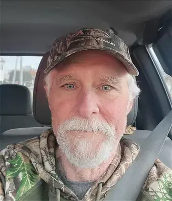 man with gray hair, beard, and mustache, wearing a camo hat and shirt, sitting in a truck wearing seatbelt