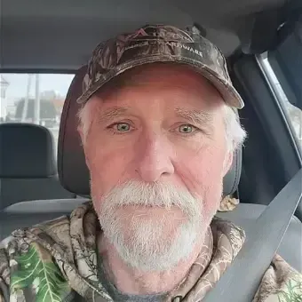 man with gray hair, beard, and mustache, wearing a camo hat and shirt, sitting in a truck wearing seatbelt