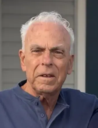Man with white hair, wearing a blue top, standing in front of a house with gray siding. 