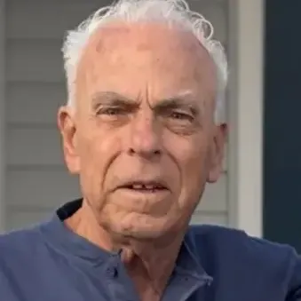 Man with white hair, wearing a blue top, standing in front of a house with gray siding. 