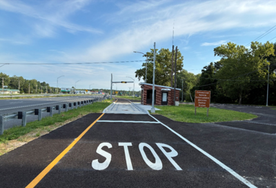 A road with, quote, "STOP" written into the lanes with white lines. Ahead, there is a scale station with a brick building and signage. The Route 13 guardrail is to the left.