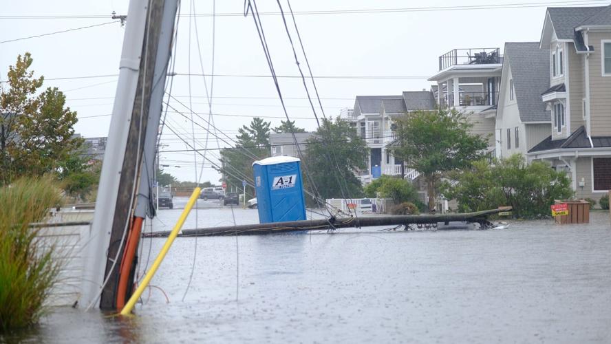 A power pole and power lines have fallen into a flooded street right next to a blue portapotty.