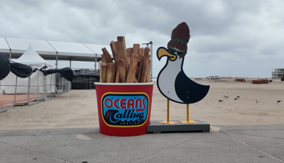 A bucket of French fries with "Oceans Calling 2026" scrawled on the front placed next to a cut-out cartoon seagull on the beach of Ocean City.