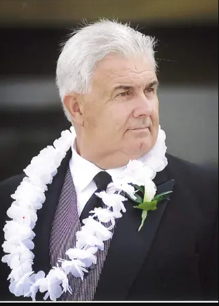 Man with white hair, wearing black jacket with white rose pin to jacket, wearing a gray vest and white lei around his neck