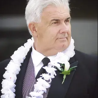 Man with white hair, wearing black jacket with white rose pin to jacket, wearing a gray vest and white lei around his neck