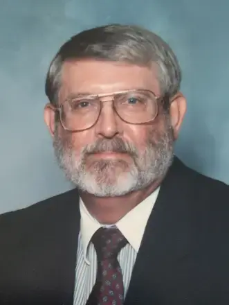 Man wearing glasses,  black suit with shirt, has salt & pepper hair short beard, and blue/gray color in the background