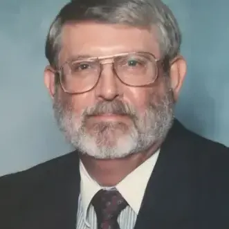 Man wearing glasses,  black suit with shirt, has salt & pepper hair short beard, and blue/gray color in the background