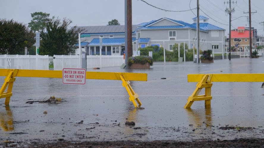 Yellow temporary fences block a road that is covered in water. One fence has a sign that says water on road, do not enter, violators fined.