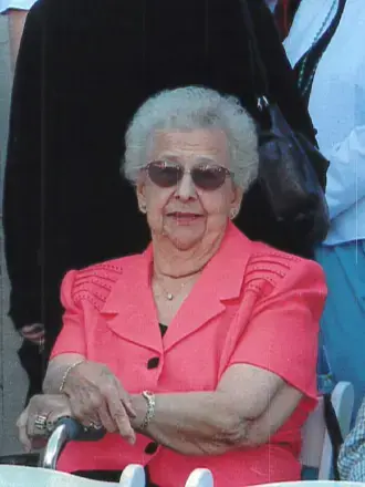 Woman with gray hair, wearing Coral color top and sunglasses. Standing on top of Statue of Liberty and can see liberty lady and harbor in background