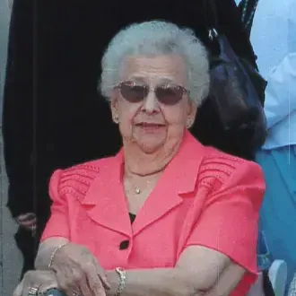 Woman with gray hair, wearing Coral color top and sunglasses. Standing on top of Statue of Liberty and can see liberty lady and harbor in background