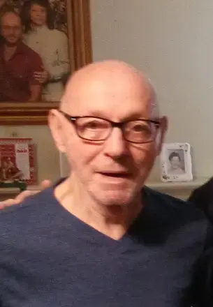 Bald man, wearing brown frame glasses and navy V-neck shirt. with framed pictures in the background