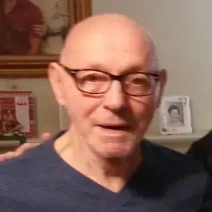 Bald man, wearing brown frame glasses and navy V-neck shirt. with framed pictures in the background