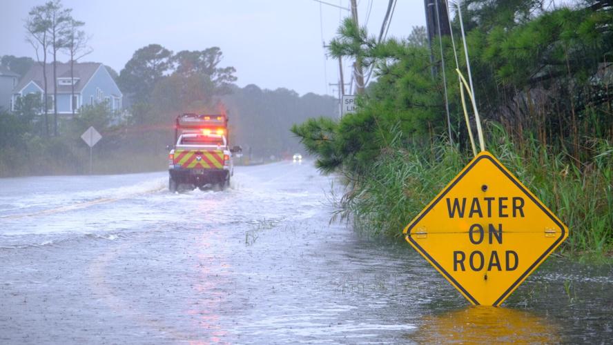 A work truck with flashing lights splashes up water on a road behind a sign that says water on road.