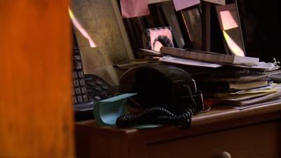 A black landline phone rests on a light brown wooden desk in what appears to be a home office surrounded by files and photographs. 