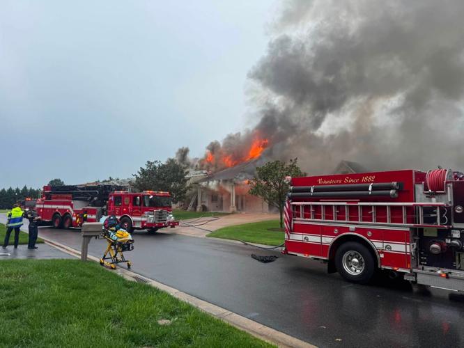 In the background, fire erupts from the roof of a brown brick home. Smoke plumes into the blue sky. On the wet road in the foreground, two fire trucks are parked, one of them has red emergency lights on. Both trucks have hoses laid out and running on the ground toward the fire. A gurney with EMS equipment is on standby, sitting on the curb of the road. Multiple first responders are standing by and observing the fire.