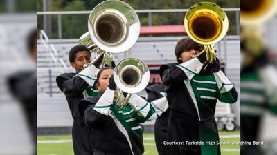 Parkside Marching Band, dressed in black, green and white uniforms, playing instruments with bleachers in the background