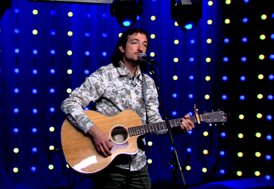 Mariano Mallia playing his guitar  singing on the MSA stage, wearing a long sleeve shirt.
