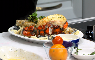 Lobster over a bed of Sea Bass Alfredo, plated on a white plate, small bowl of an orange and small tomato in front of plated entrée. 