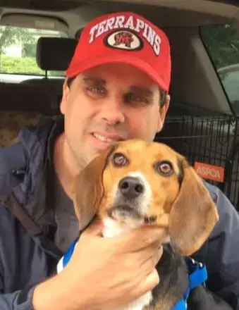 Man holding a beagle puppy, wearing a red MD Terps hat and blue jacket sitting in a car 