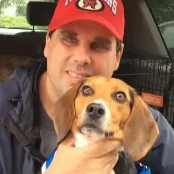 Man holding a beagle puppy, wearing a red MD Terps hat and blue jacket sitting in a car 