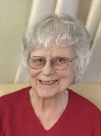 woman with short white hair, wearing glasses and red B-neck top sitting on a cream color seat and white drapes in the background