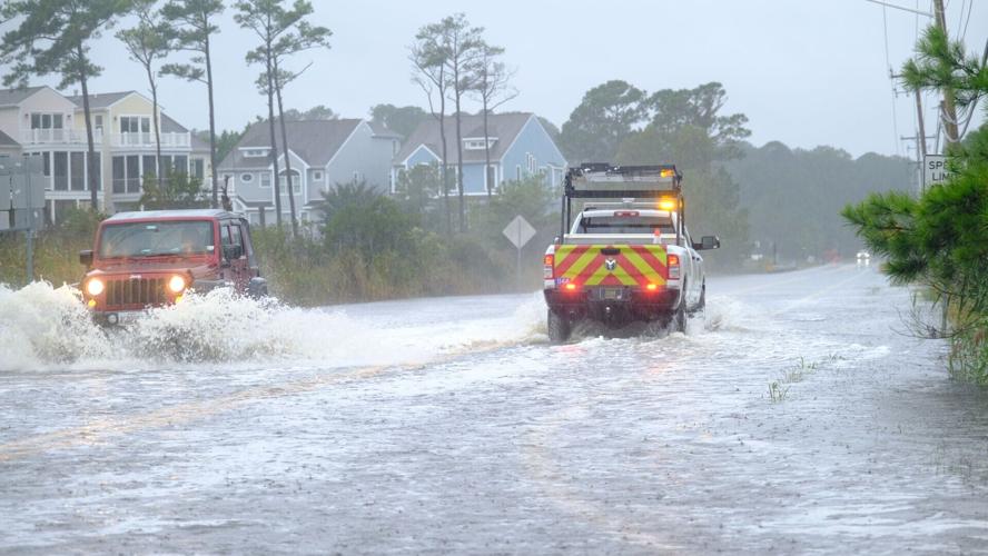A work truck passes a red jeep as both drive through high water, splashing.