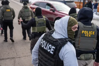 An ICE agent in a mask turns around as five other agents walk ahead of him.